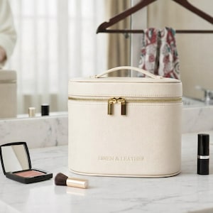 May include: A cream-coloured, oval-shaped vanity case with a top handle and gold-tone zip closure. The front of the case features the embossed text LINEN & LEATHER. It is positioned on a marble surface alongside a compact mirror and bottles.