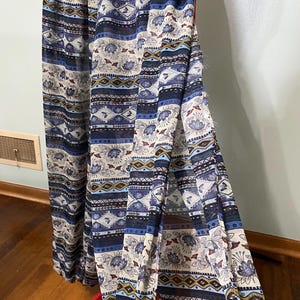 Könnte beinhalten: Ein bodenlanger Rock aus leichtem, transparentem Stoff mit einem mehrfarbigen Muster aus blauen, weißen und goldenen Blumen- und Geometriestreifen. Das Kleidungsstück verfügt über einen hohen Seitenschlitz, der den Lagenaufbau des Stoffes zeigt.