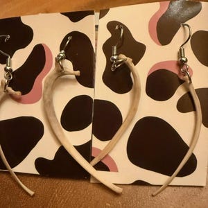 May include: A pair of unique earrings made from curved, light-colored bone pieces. The earrings hang from silver-colored hooks and are displayed on a card with a black and pink cow-print pattern.