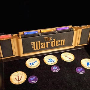 Puede incluir: Una caja de juego de mesa con el título "The Warden" en letras doradas. La caja está abierta, revelando piezas de juego y ranuras para cartas. Varias fichas circulares con ilustraciones están dispersas sobre la superficie de juego.