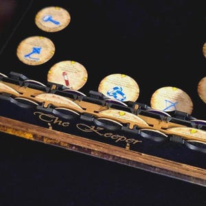 May include: Wooden board game component with the text "The Keeper" and several slots. The component is tan and black with a skull icon. Several circular game pieces with symbols are also visible.
