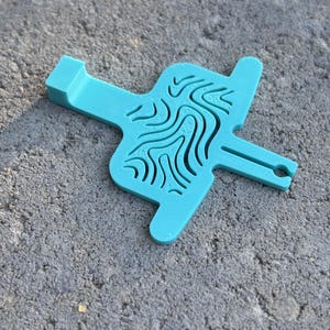 Puede incluir: Una herramienta plana de color verde azulado con varias puntas, que presenta una sección central con un patrón de mapa topográfico en relieve. El objeto tiene una lengüeta rectangular en un extremo y un brazo estrecho ranurado con una abertura.
