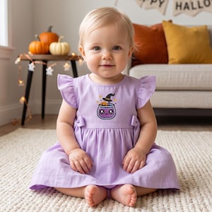 May include: A light purple dress with flutter sleeves and a gathered waist, featuring a central graphic of a smiling purple potion cauldron wearing a black witch hat with small yellow stars surrounding the design on the chest area.