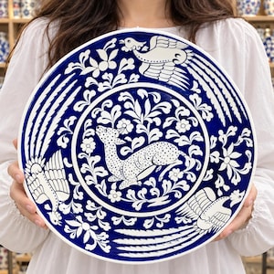 Könnte beinhalten: Ein runder Keramikteller mit tiefblauem Hintergrund und handgemalten weißen Mustern. In der Mitte ist ein gepunktetes Reh umgeben von Blumenmotiven abgebildet, während der äußere Rand drei stilisierte Vögel mit langen Schwanzfedern zeigt.