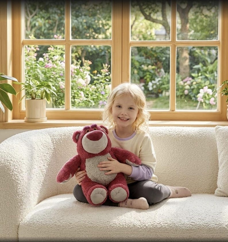 May include: A young girl smiles while holding a plush, red teddy bear with a light brown belly. The bear has a friendly expression. The girl is sitting on a white, textured couch in front of a window.