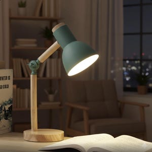Puede incluir: Una lámpara de escritorio verde con base y brazo de madera. La pantalla de la lámpara está dirigida hacia un libro abierto. La lámpara está sobre un escritorio de madera. El fondo incluye una estantería y una ventana.