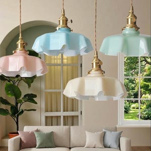 May include: Four pendant lights with ruffled, pastel-coloured shades hang from the ceiling. The shades are in shades of pink, blue, green, and cream. The fixtures have brass-coloured hardware and are suspended above a beige sofa with throw cushions.