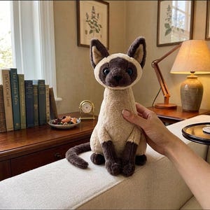 May include: A plush Siamese cat toy with a beige body, dark brown ears, face, paws, and tail. The cat has blue eyes and is sitting upright. The toy is being held on a white couch. The background includes a wooden table, books, and a lamp.