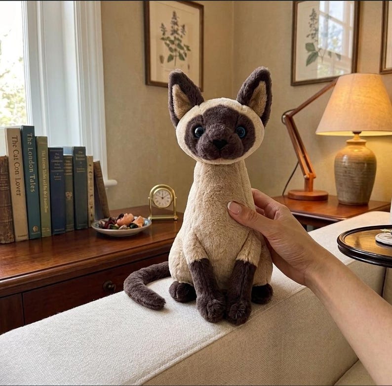 May include: A plush Siamese cat toy with a beige body, dark brown ears, face, paws, and tail. The cat has blue eyes and is sitting upright. The toy is being held on a white couch. The background includes a wooden table, books, and a lamp.