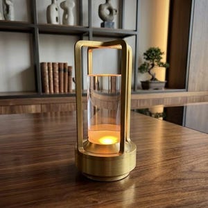 May include: A gold-colored lantern with a cylindrical glass insert, illuminated by a warm light. The lantern has a square top and base, and is sitting on a dark wooden surface. Decorative items are visible in the background.
