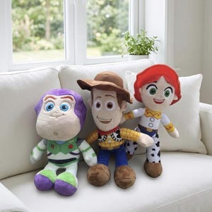 May include: Three plush toy figures of characters from the animated film series Toy Story. Buzz Lightyear is on the left, Woody in the center, and Jessie on the right. The toys are on a white couch in front of a window.
