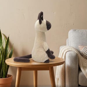 May include: A plush Siamese cat stuffed animal with cream-colored fur and dark brown accents on its ears, face, paws, and tail. The stuffed animal is sitting on a round wooden table. The text "FULL FILLING" is at the top of the image.