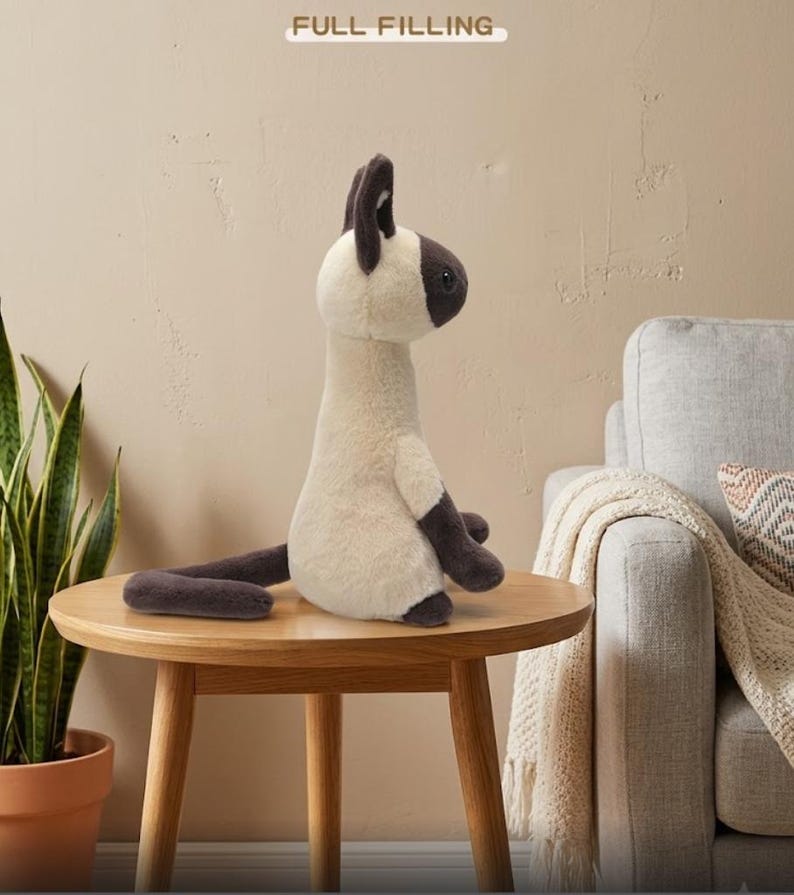 May include: A plush Siamese cat stuffed animal with cream-colored fur and dark brown accents on its ears, face, paws, and tail. The stuffed animal is sitting on a round wooden table. The text "FULL FILLING" is at the top of the image.