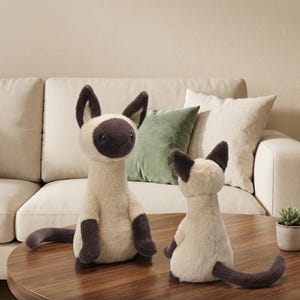 May include: Two plush Siamese cat stuffed animals. The cats have cream-colored bodies, dark brown ears, paws, and tails. One cat is upright, and the other is seated. They are on a wooden table in front of a beige sofa.
