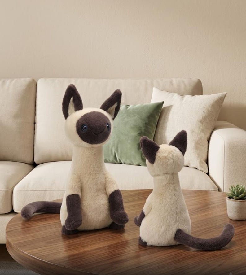 May include: Two plush Siamese cat stuffed animals. The cats have cream-colored bodies, dark brown ears, paws, and tails. One cat is upright, and the other is seated. They are on a wooden table in front of a beige sofa.