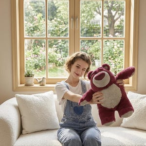 May include: A plush, burgundy-colored teddy bear with a light tan belly and a smiling face. The bear is being held up in front of a window with a garden view. The bear has outstretched arms.