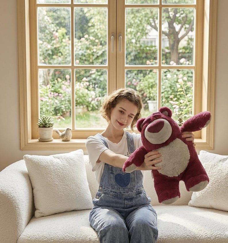May include: A plush, burgundy-colored teddy bear with a light tan belly and a smiling face. The bear is being held up in front of a window with a garden view. The bear has outstretched arms.