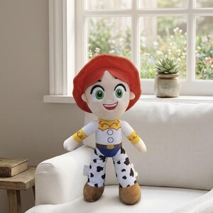 May include: A soft toy doll featuring a character with red hair, a white shirt with yellow details, blue jeans, and black and white spotted trousers. The doll has green eyes and a smiling face. It stands on a white sofa near a window.