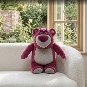 May include: A plush, pink teddy bear with a heart-shaped nose and a grumpy expression. The bear has a white belly and is sitting on a white, textured couch. The background includes a window and greenery.