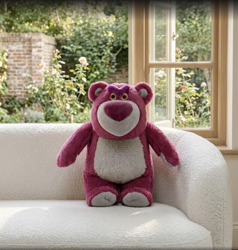 May include: A plush, pink teddy bear with a heart-shaped nose and a grumpy expression. The bear has a white belly and is sitting on a white, textured couch. The background includes a window and greenery.