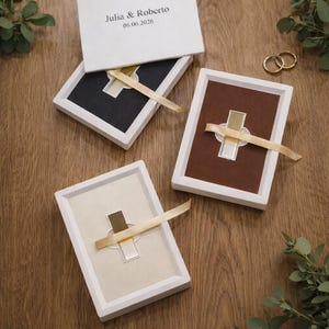 Op de afbeelding: Trouwringdozen met witte frames en gekleurde interieurs: zwart, bruin en crème. Elke doos heeft een gouden kruisvormig accent en een crèmekleurig lint. De bovenste doos is open en toont de namen "Julia & Roberto" en de datum "06.06.2026".