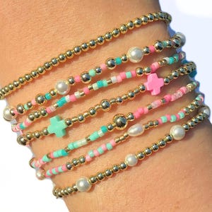 May include: A stack of seven beaded bracelets featuring gold-tone metallic spheres, small white pearls, and colorful seed beads in shades of pink and turquoise. The bracelets include two small cross-shaped charms, one in pink and one in turquoise.