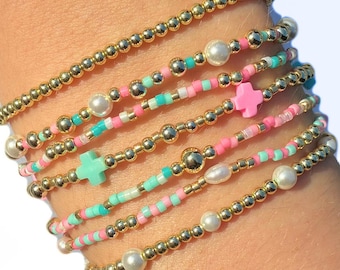 Può includere: Una pila di sette braccialetti di perline con sfere metalliche dorate, piccole perle bianche e perline colorate nelle tonalità del rosa e del turchese. I braccialetti includono due piccoli ciondoli a forma di croce, uno rosa e uno turchese.