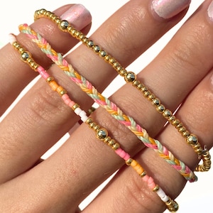 May include: Close-up of four colorful bracelets. Two bracelets feature gold beads, while the other two have braided and beaded designs in pastel pink, orange, yellow, and green. The bracelets are delicate and appear handmade.