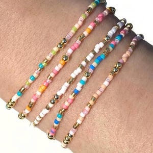 Könnte beinhalten: Fünf zarte Perlenarmbänder mit kleinen, bunten Glasperlen in Rosa, Blau, Orange, Grün und Weiß, durchsetzt mit kleinen, runden, metallisch goldfarbenen Abstandsperlen, die in einem sich wiederholenden Muster angeordnet sind.