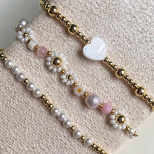 May include: Close-up of three beaded bracelets. One bracelet features white pearls and gold beads. Another has daisy and pearl accents, with pink and white beads. The third bracelet has a white heart charm and gold beads.