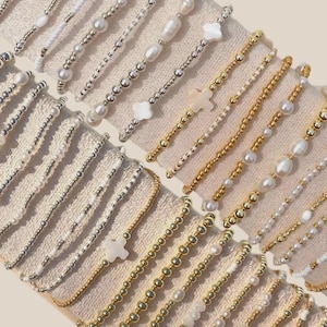 以下が含まれることがあります： 金、銀、白のビーズブレスレットの詰め合わせ。ブレスレットには、パール、小さなビーズ、十字架や花の形などの装飾的なチャームが付いています。ブレスレットはニュートラルカラーの生地の上に配置されています。