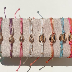 May include: A collection of colorful seashell charm bracelets. Each bracelet features a gold-colored seashell pendant and is strung on a cord in various colors, including pink, purple, white, and blue. The bracelets are adorned with small gold beads.