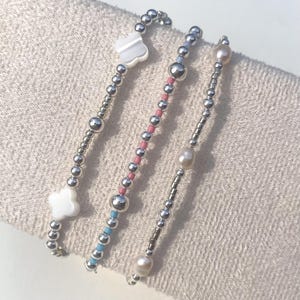 May include: Three delicate bracelets with silver beads and accents. One features a white clover charm, another has pink and blue beads, and the third showcases pearls. The bracelets are displayed on a light-colored fabric.