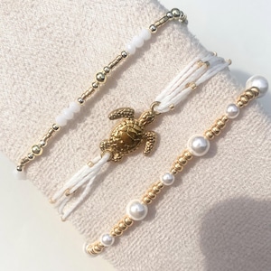 Puede incluir: Tres pulseras con cuentas. Una es blanca con cuentas doradas, otra tiene un dije de tortuga dorada y la tercera presenta perlas y cuentas doradas. Las pulseras se exhiben sobre una tela blanca.