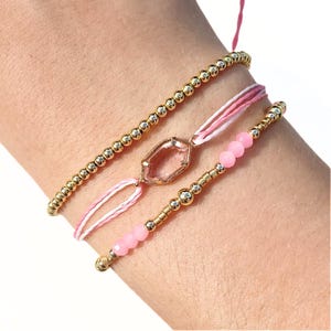 Puede incluir: Se muestran tres pulseras en una muñeca. La pulsera superior es de cuentas doradas. La pulsera del medio tiene hilo rosa y blanco con una gema rosa. La pulsera inferior tiene cuentas rosas y cuentas doradas. Las pulseras son delicadas y femeninas.