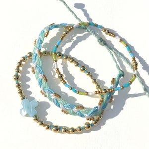 May include: Four bracelets in shades of blue, white, and gold. One bracelet features a light blue cross-shaped charm. The other bracelets have gold beads and braided or beaded designs. The bracelets are on a white surface.