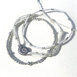 Puede incluir: Tres pulseras de cuentas blancas y plateadas. Una pulsera presenta un dije de amonita plateada. Las pulseras están hechas con cordón blanco y cuentas plateadas de varios tamaños. Las pulseras están sobre un fondo blanco.