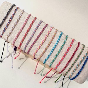 Puede incluir: Una colección de pulseras de la amistad trenzadas en varios colores, incluyendo negro, rojo, blanco, rosa, morado, amarillo, azul y verde. Cada pulsera se muestra sobre una superficie de tela beige, mostrando los intrincados patrones de trenzado y los tonos vibrantes.