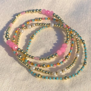 Puede incluir: Conjunto de pulseras de cuentas de colores. Las pulseras presentan cuentas doradas, gemas rosas, perlas blancas y pequeñas cuentas de colores en tonos azules, naranjas y rosas. Las pulseras están dispuestas sobre una superficie blanca.