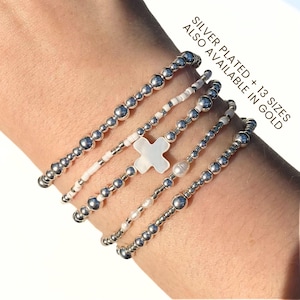 May include: Silver-plated beaded bracelets in various designs. One bracelet features a white cross, another has a pearl, and others have small white beads. The text in the image says "Silver Plated + 13 Sizes Also Available in Gold."
