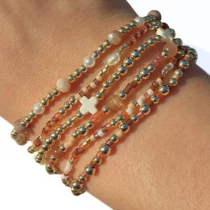 Puede incluir: Una pila de cinco pulseras de cuentas que presenta una mezcla de esferas doradas metálicas, perlas lisas de color crema, cuentas translúcidas en tonos melocotón y pequeñas cuentas de semillas marrones. Una pulsera incluye una cuenta blanca.