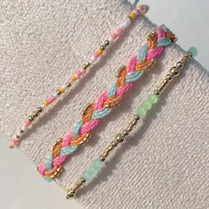 Könnte beinhalten: Drei farbenfrohe Armbänder. Eines ist ein geflochtenes Design mit rosa, orangefarbenen und blauen Fäden, akzentuiert mit goldenen Perlen. Ein weiteres ist ein Perlenarmband mit rosa, gelben und weißen Perlen. Das dritte Armband hat grüne Perlen und goldene Akzente.