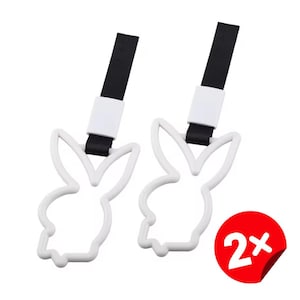 May include: Two white bunny-shaped car accessories with black straps and white clasps. The bunny outline is a recognizable logo. A red sticker with the number "2x" indicates a set of two items.
