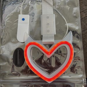 May include: A heart-shaped LED light with a white frame and a bright orange outline. The light is attached to a white strap and a small white control box. The product is packaged in a clear plastic bag.