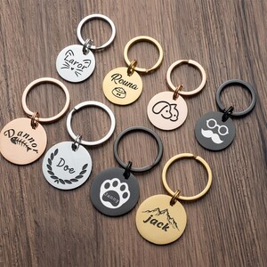 May include: A collection of personalized pet ID tags and keychains in various metallic finishes. The tags feature engraved designs such as a cat face, paw print, and mountain scene, along with names and other details. Each tag is attached to a metal ring.