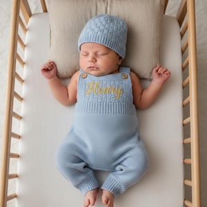 May include: A light blue knitted romper and matching beanie set featuring a textured basketweave pattern on the chest and hat. The romper has two small wooden buttons at the shoulders and the name Henry embroidered in gold cursive script.
