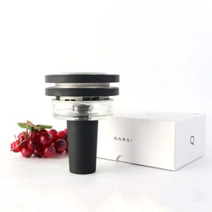 May include: A contemporary wine bottle stopper in black and clear. The stopper features a black base and top with a clear glass section. A white box with the text "RAAS²" and a cluster of red grapes are also visible.