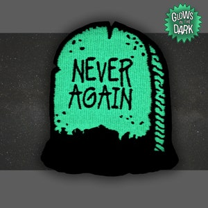 May include: Green glow-in-the-dark embroidered patch shaped like a tombstone with the text "NEVER AGAIN" in black.