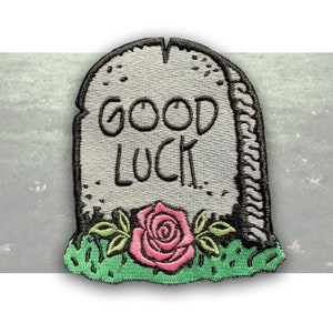 May include: Embroidered iron-on patch featuring a gray tombstone with the words "GOOD LUCK" in black lettering. A pink rose with green leaves is at the base of the tombstone.