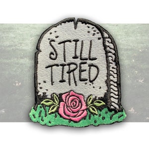 May include: Embroidered iron-on patch featuring a tombstone with the text "Still Tired" and a pink rose with green leaves.